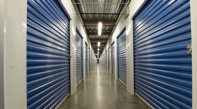 A long well lit hallway of indoor storage units with blue doors