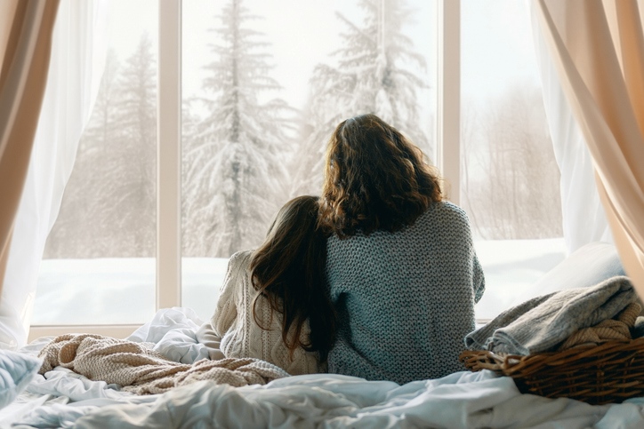 A mother and daughter watch the snow fall from a window.